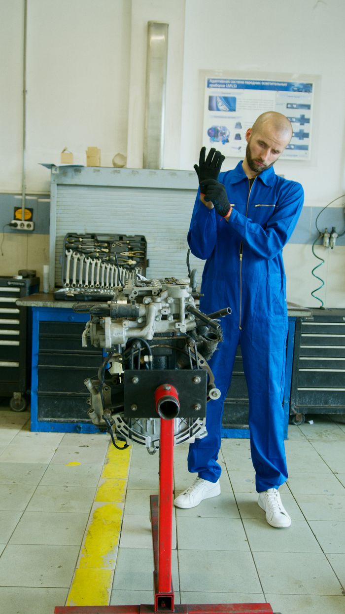 Mechanic in blue overalls adjusting gloves by engine in garage setting.